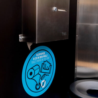Toilettenpapierspender aus Edelstahl in der Libre Trockentoilette mit blauem Hinweisschild.