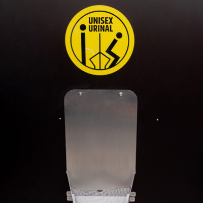 Edelstahl-Unisexurinal in der Libre Trockentoilette vor schwarzer Wand mit gelbem Hinweisschild: Unisex Urinal