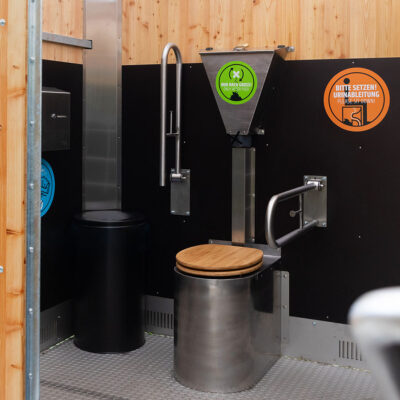 Innenansicht der Libre Trockentoilette mit Edelstahltoilette Peepot und Holzsitz, Streuspender Spencer mit grünem Hinweis, Klappstützgriffe und orangem Hinweisschild.