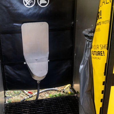 Innenansicht des Unisexurinals mit Edelstahlbecken, schwarzer Rückwand und Müllbeutelhalterung neben gelber Türplane.