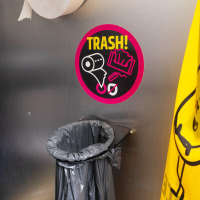 Innenansicht des Unisexurinals Unipee mit rundem Trash-Hinweis-Sticker für Papierabfall und schwarzem Müllbeutel an Metallwand.