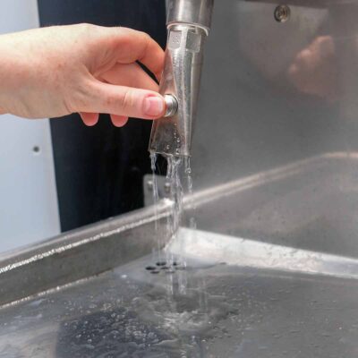 Detailansicht einer Hand, die den Wasserhahn der Libre-DIN Trockentoilette betätigt, mit fließendem Wasser.