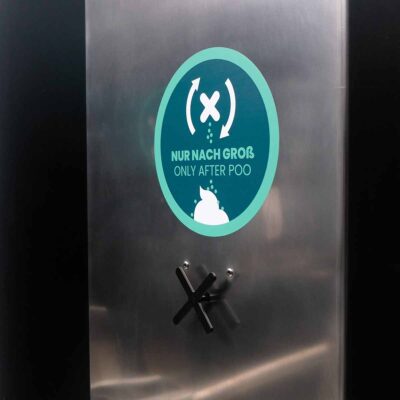 Detailansicht der Edelstahlfläche des Streuspenders Spencer mit Hinweisschild mit Text „Nur nach Groß – / Only after poo“ und Symbol für Einstreu-Nutzung nach dem Toilettengang.