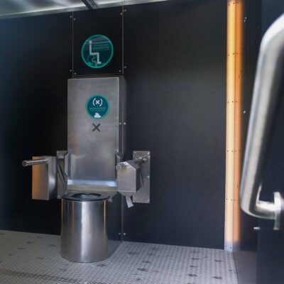 Innenansicht Libre DIN barrierefreie, geschlechterneutrale Trockentoilette mit Edelstahltoilette und Klappstützgriffen an schwarzer Wand.