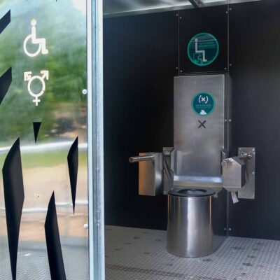 Innenansicht Libre DIN barrierefreie, geschlechterneutrale Trockentoilette mit Edelstahltoilette und Klappstützgriffen an schwarzer Wand.
