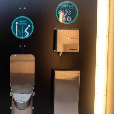Innenansicht Humero Trockentoilette mit Unisexurinal aus Edelstahl, Papierhandtuchspender und Abfallbehälter an schwarzer Wand.