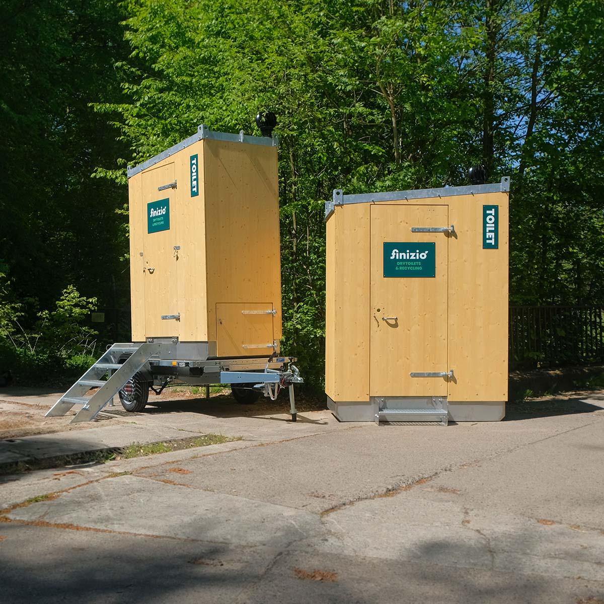 Außenansicht Humero Rollin’ auf Trailer links und Humero Trockentoilette rechts vor grünem Wald