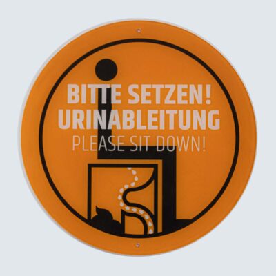 Rundes orangefarbenes Schild mit schwarzem Piktogramm und Text „Bitte setzen! / Please sit down!“.