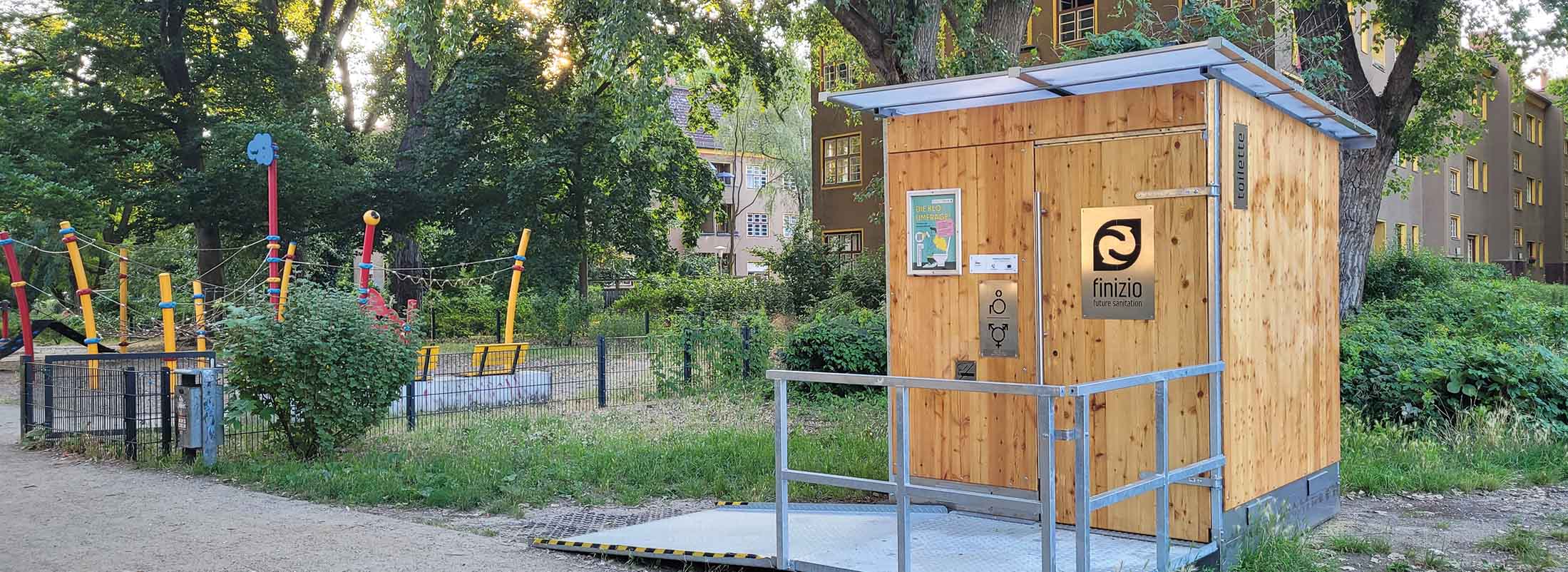 Libre-Light-Finizio-Berliner-Parktoilettenprojekt Ein öffentliches Trockentoiletten-Häuschen von Finizio vor einem urbanen Spielplatz umgeben von Bäumen.
