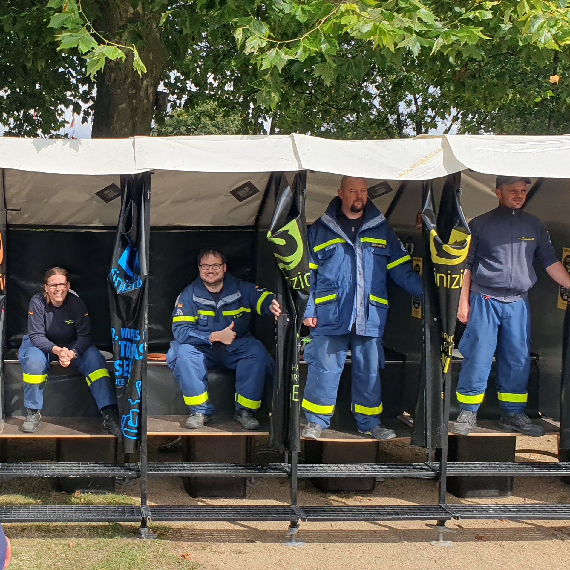 Finizio_THW_Aufbau_Humusphaere_mobile_Toilette_Katastrophenschutz Vier Menschen die die Uniform des technischen Hilfswerks tragen, stehen und sitzen in geöffneten Toilettenkabinen.